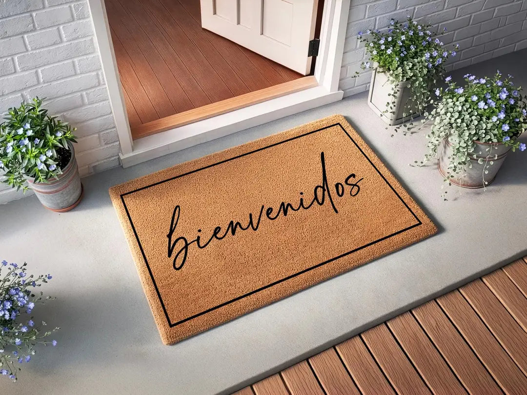 Coir doormat with a framed bienvenidos script message at an open white door with white brick siding and potted blue flowers