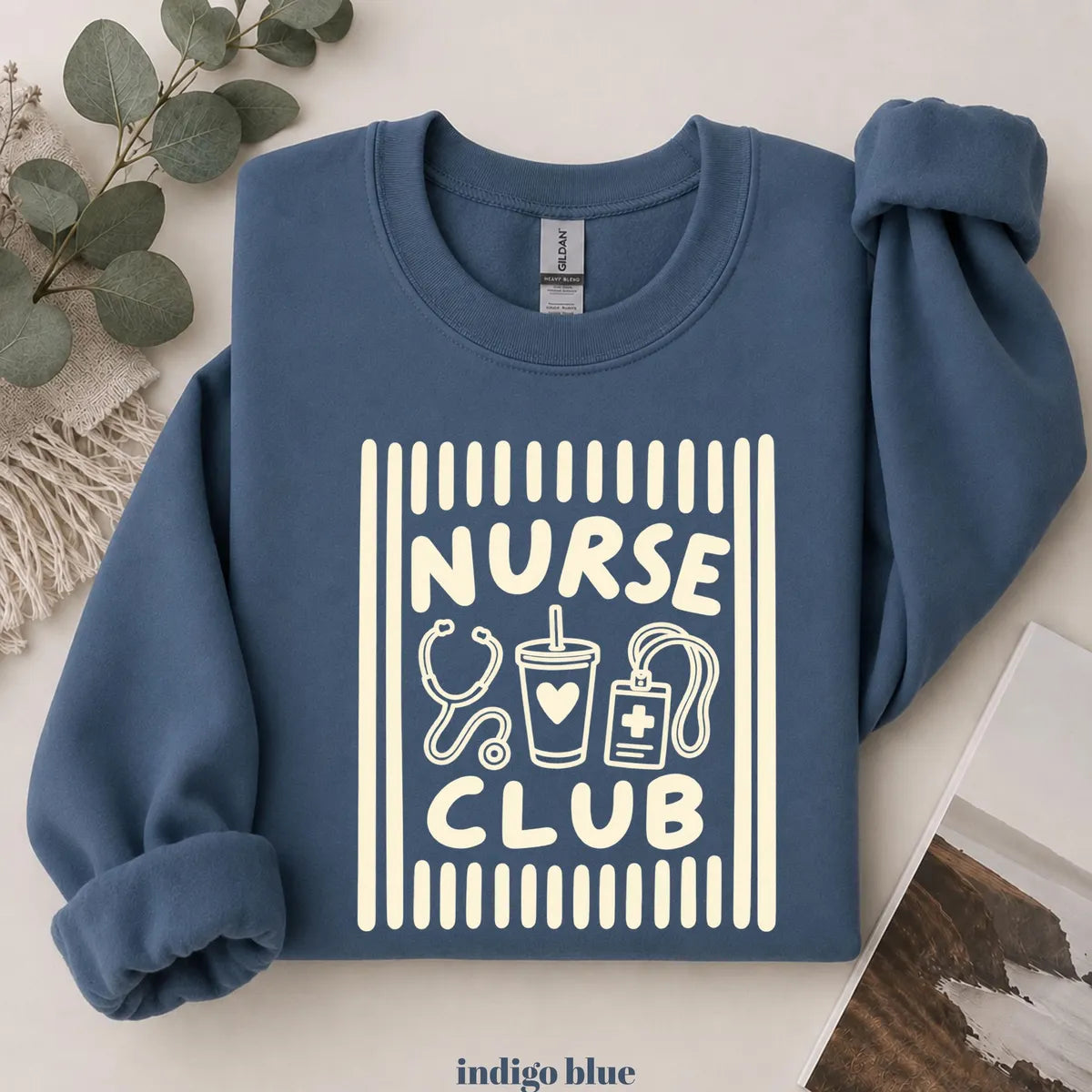 Indigo Blue crewneck sweatshirt with printed a retro striped frame with a stethoscope, coffee cup and badge above the words nurse club, flat lay view.