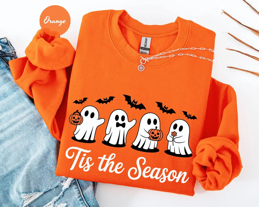 Orange crewneck sweatshirt with printed four cute ghosts with pumpkins and bats above the words tis the season, flat lay view.