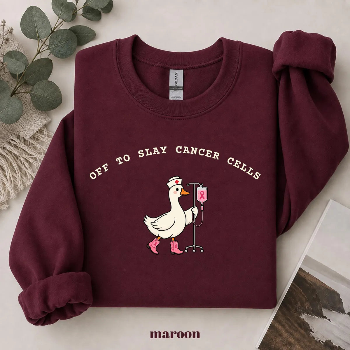 Maroon crewneck sweatshirt with printed a nurse goose in pink boots with an IV pole and the words off to slay cancer cells, flat lay view.