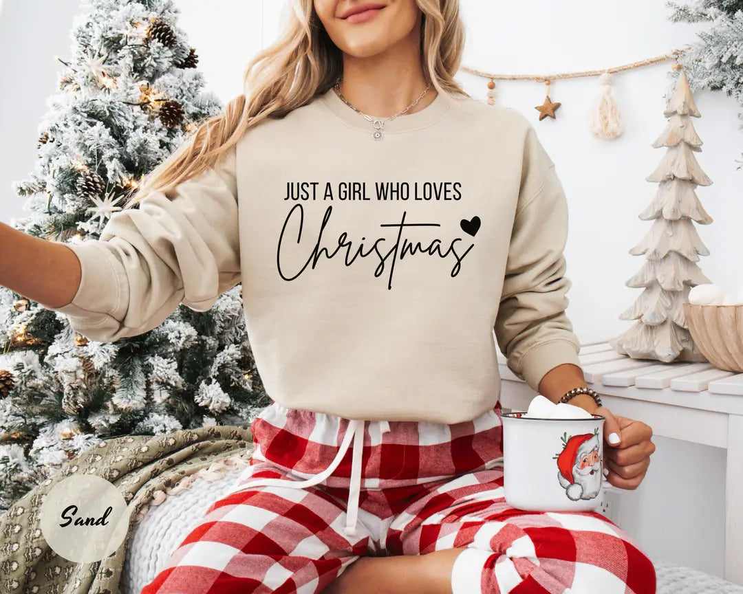 Sand crewneck sweatshirt with script lettering that reads just a girl who loves Christmas with a small heart, worn by a model.