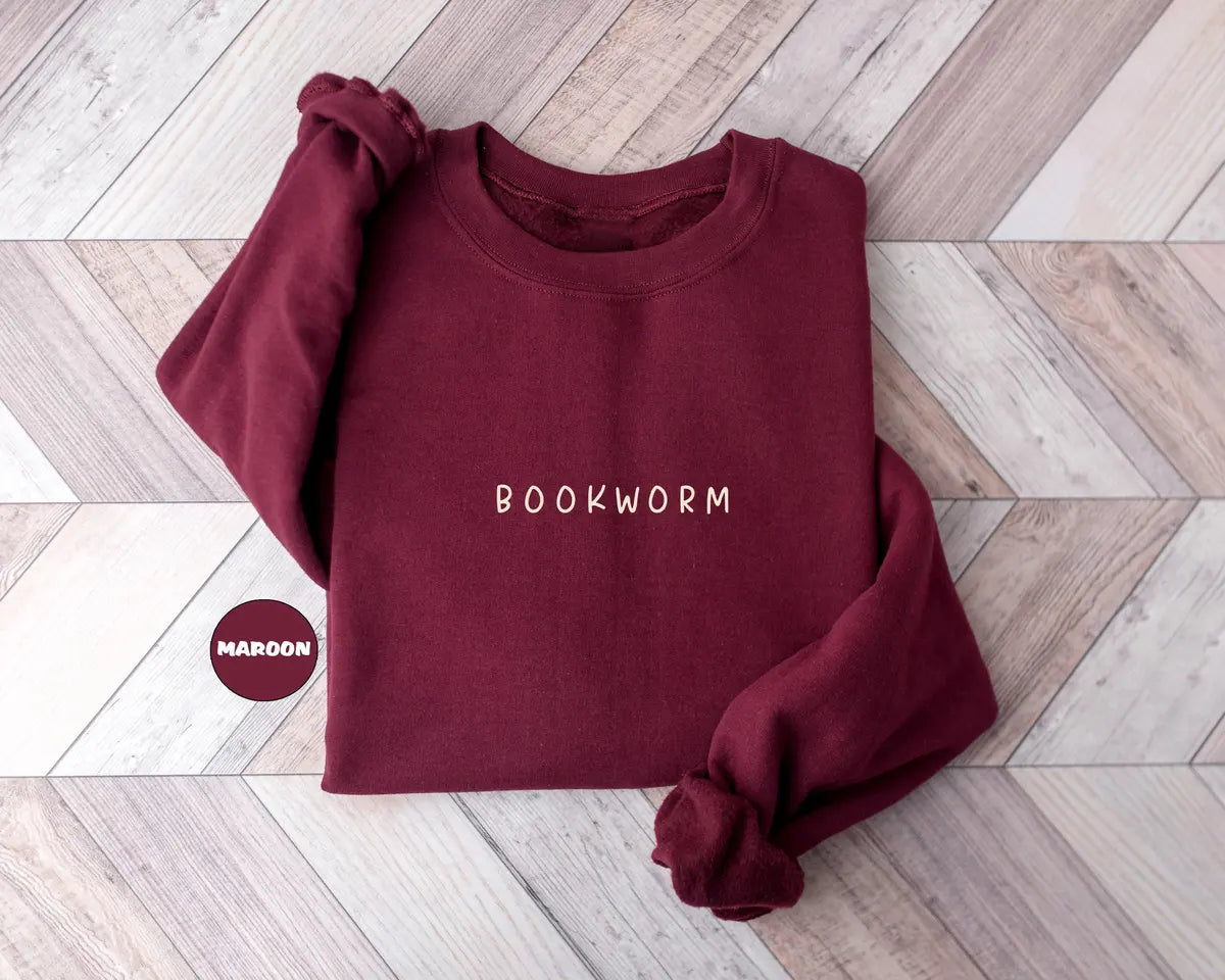 Maroon crewneck sweatshirt with small minimal lettering that reads bookworm on the chest, flat lay view.