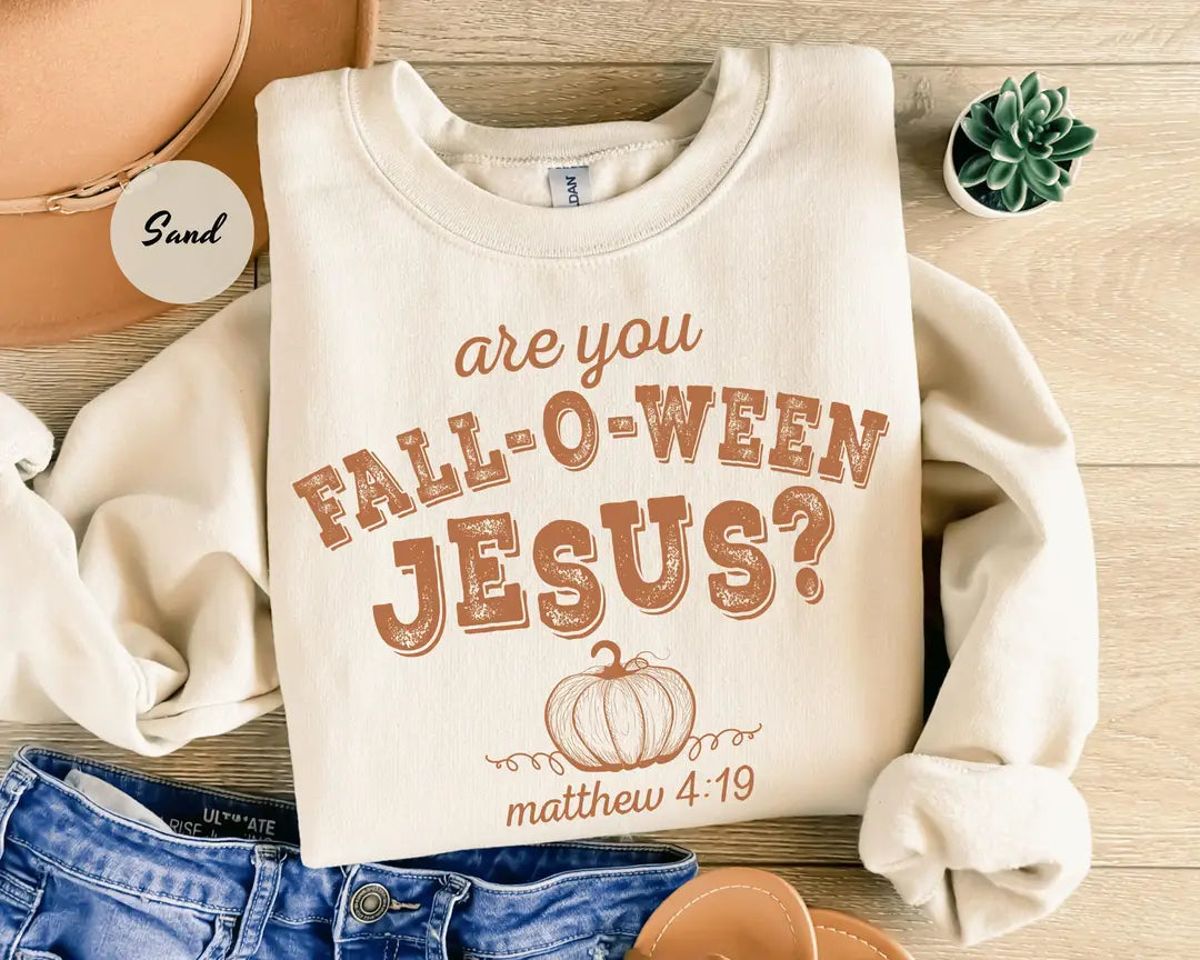 Sand crewneck sweatshirt with printed a pumpkin with the words are you fall-o-ween jesus and matthew 4:19 in a western style font, flat lay view.
