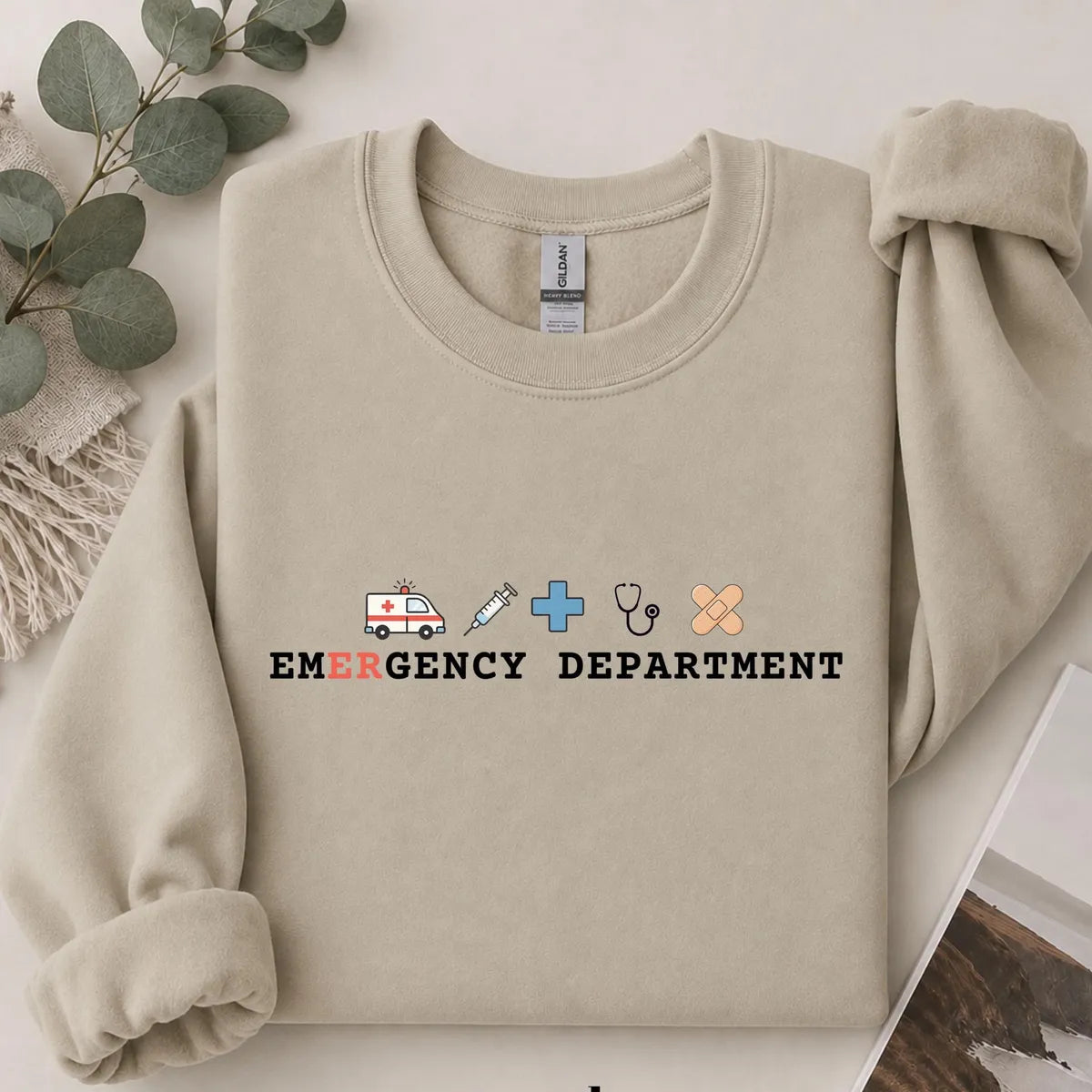Sand crewneck sweatshirt with printed an ambulance, syringe, cross, stethoscope and bandage above the words emergency department, flat lay view.