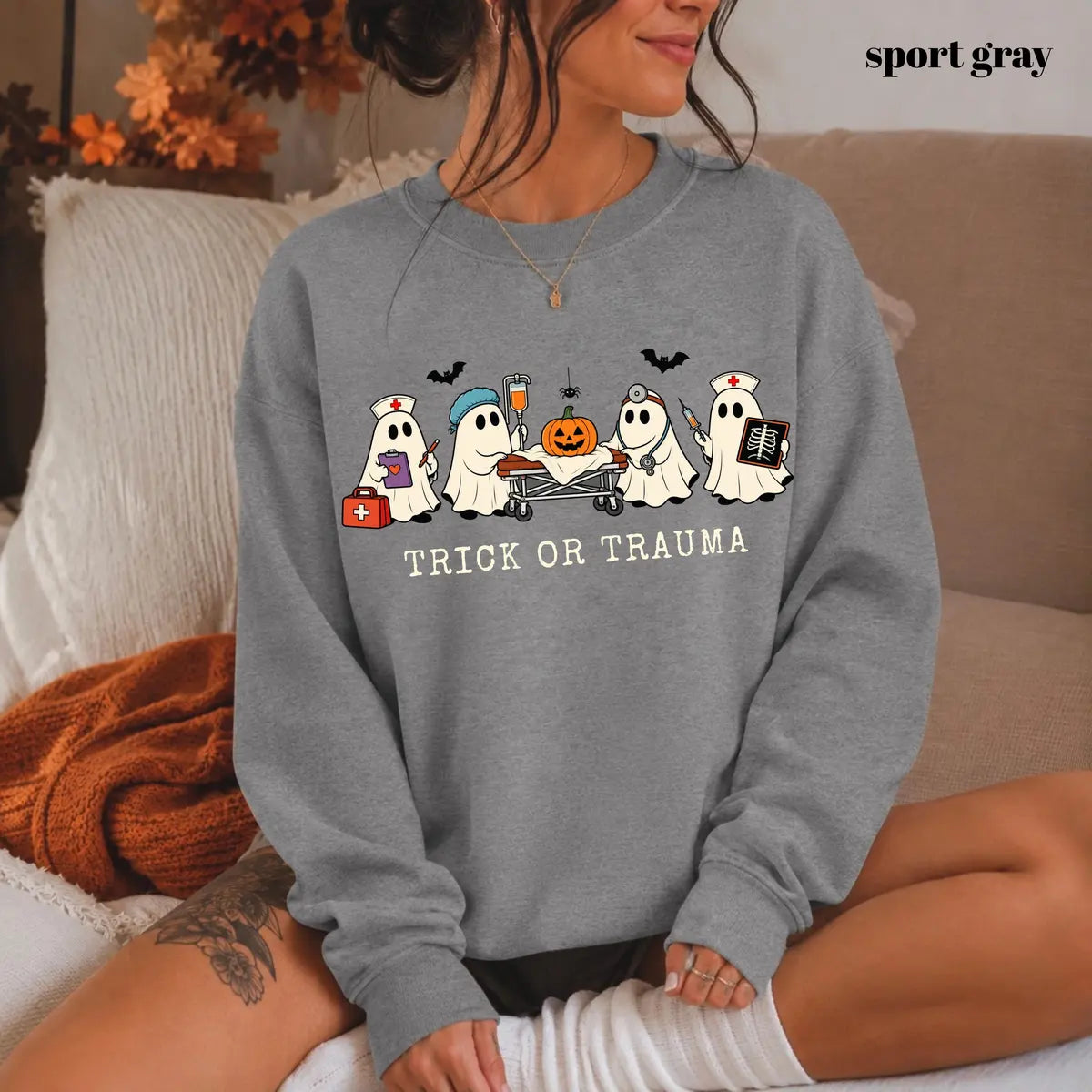 Sport Grey crewneck sweatshirt with printed nurse ghosts with a pumpkin and the words trick or trauma, worn by a model.