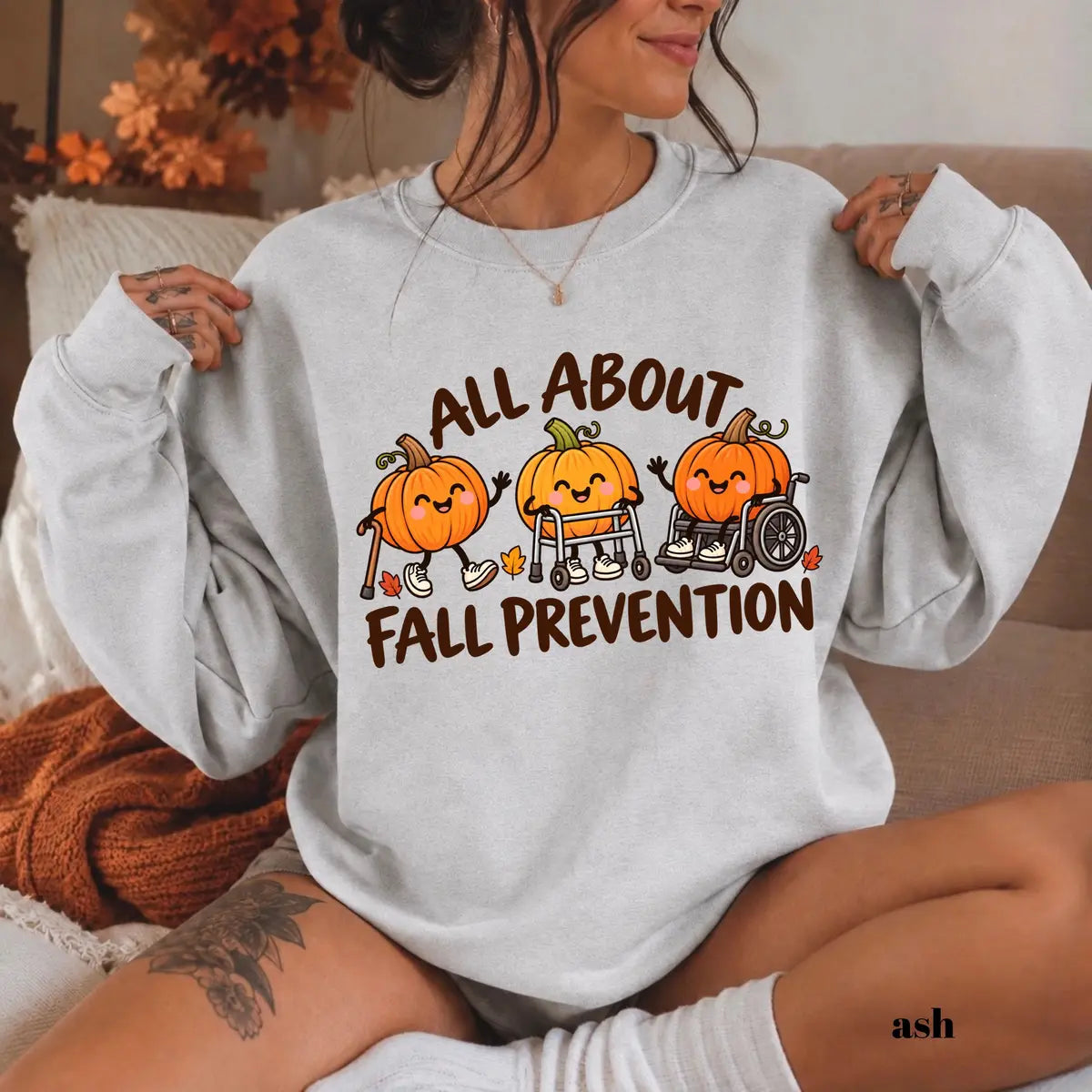 Ash crewneck sweatshirt with printed pumpkins with a walker and wheelchair above the words all about fall prevention, worn by a model.