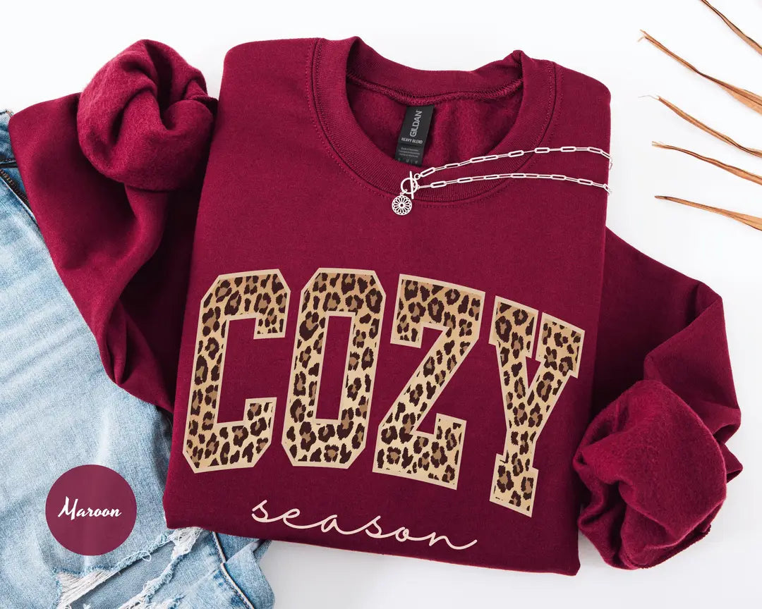 Maroon crewneck sweatshirt with bold leopard print cozy lettering above the script words season, flat lay view.