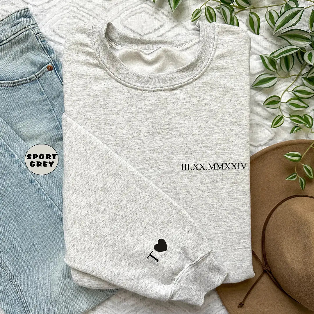 Sport Grey crewneck sweatshirt with a custom date in Roman numerals on the chest and an initial with a heart on the cuff, flat lay view.