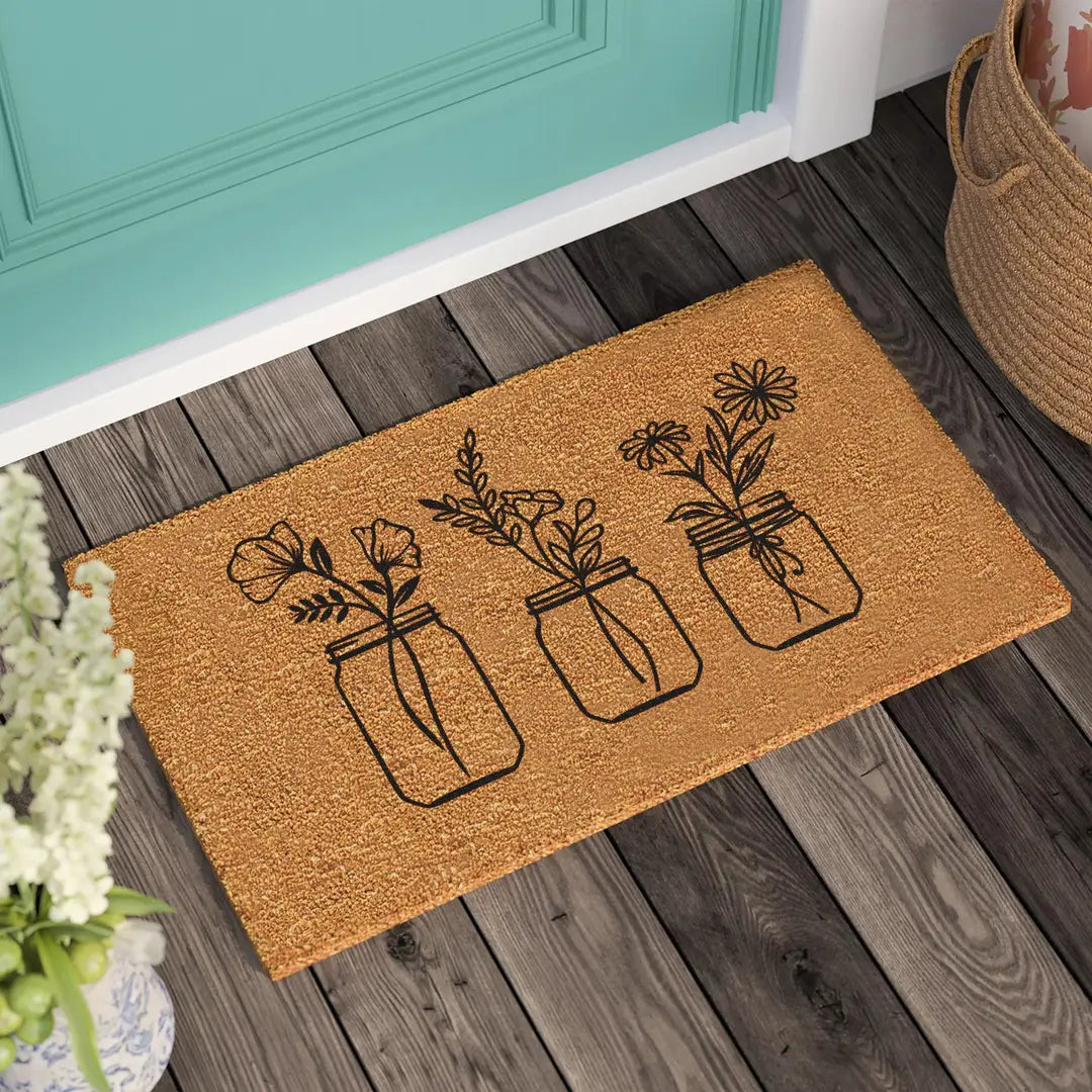 Coir doormat with three line art wildflower bouquets in jars on a wooden deck in front of a teal door