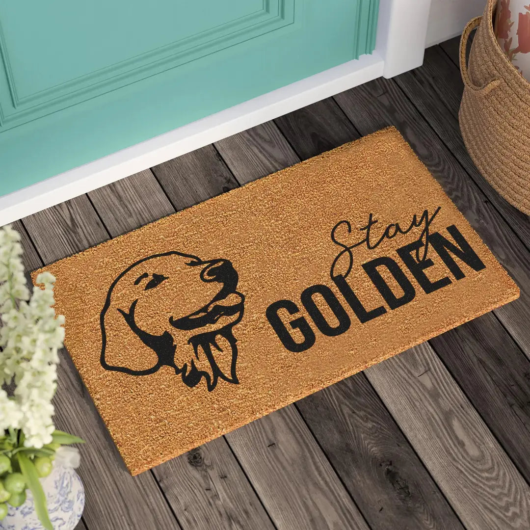 Coir doormat with a line-art golden retriever head and Stay Golden lettering on a wooden porch beside a teal door and woven basket