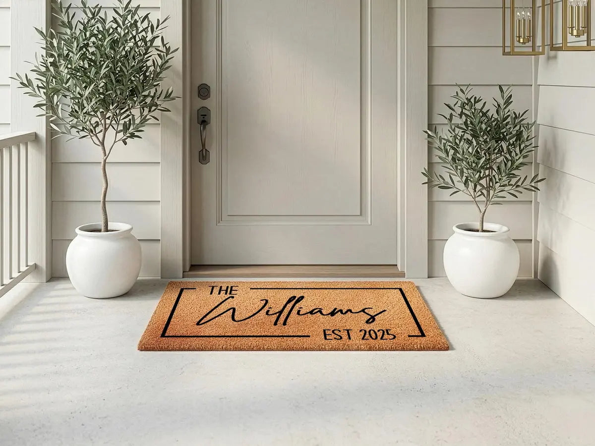 Coir doormat with a framed family name and established year at a white door flanked by potted olive trees