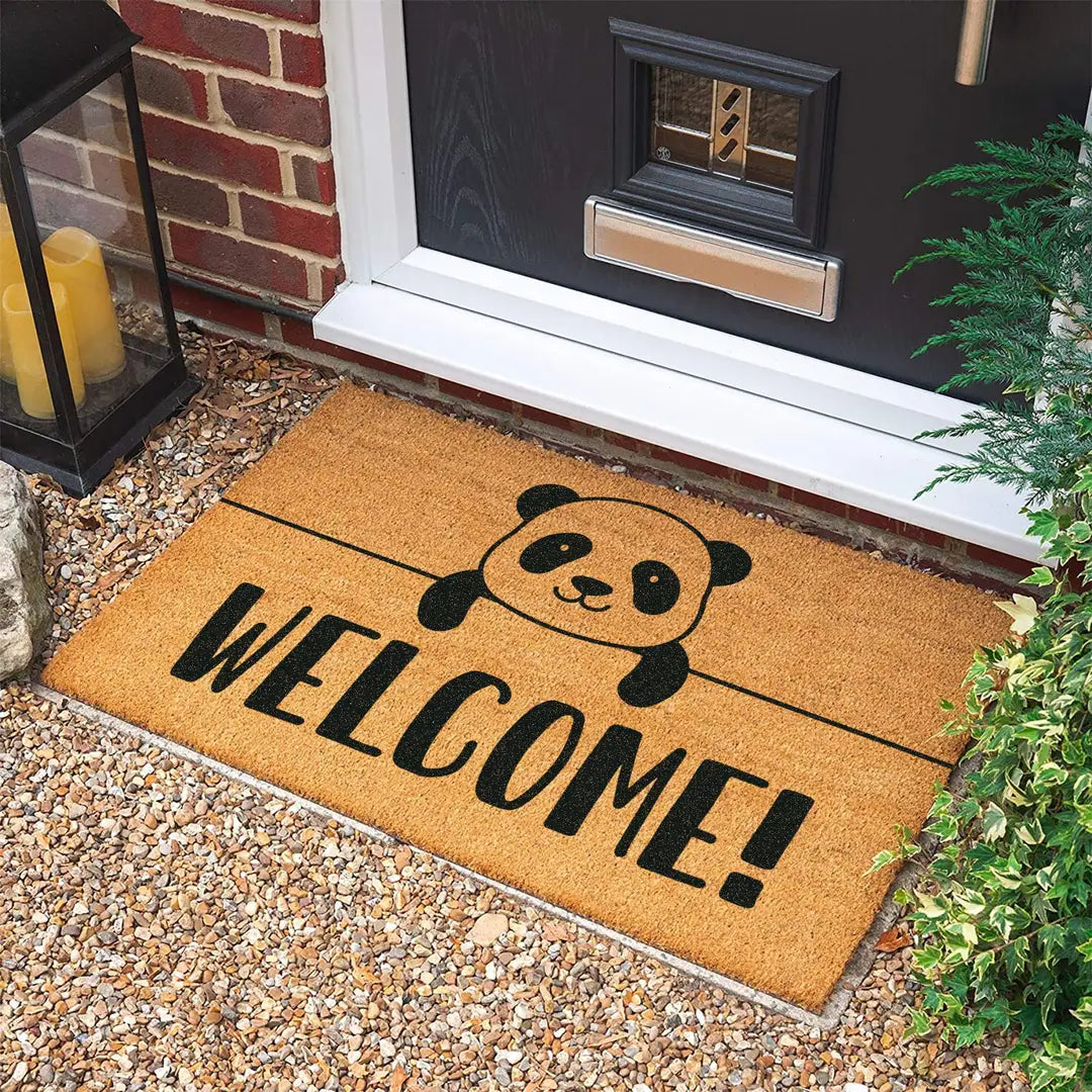 Coir doormat with a peekaboo panda and bold WELCOME lettering at a dark front door on gravel