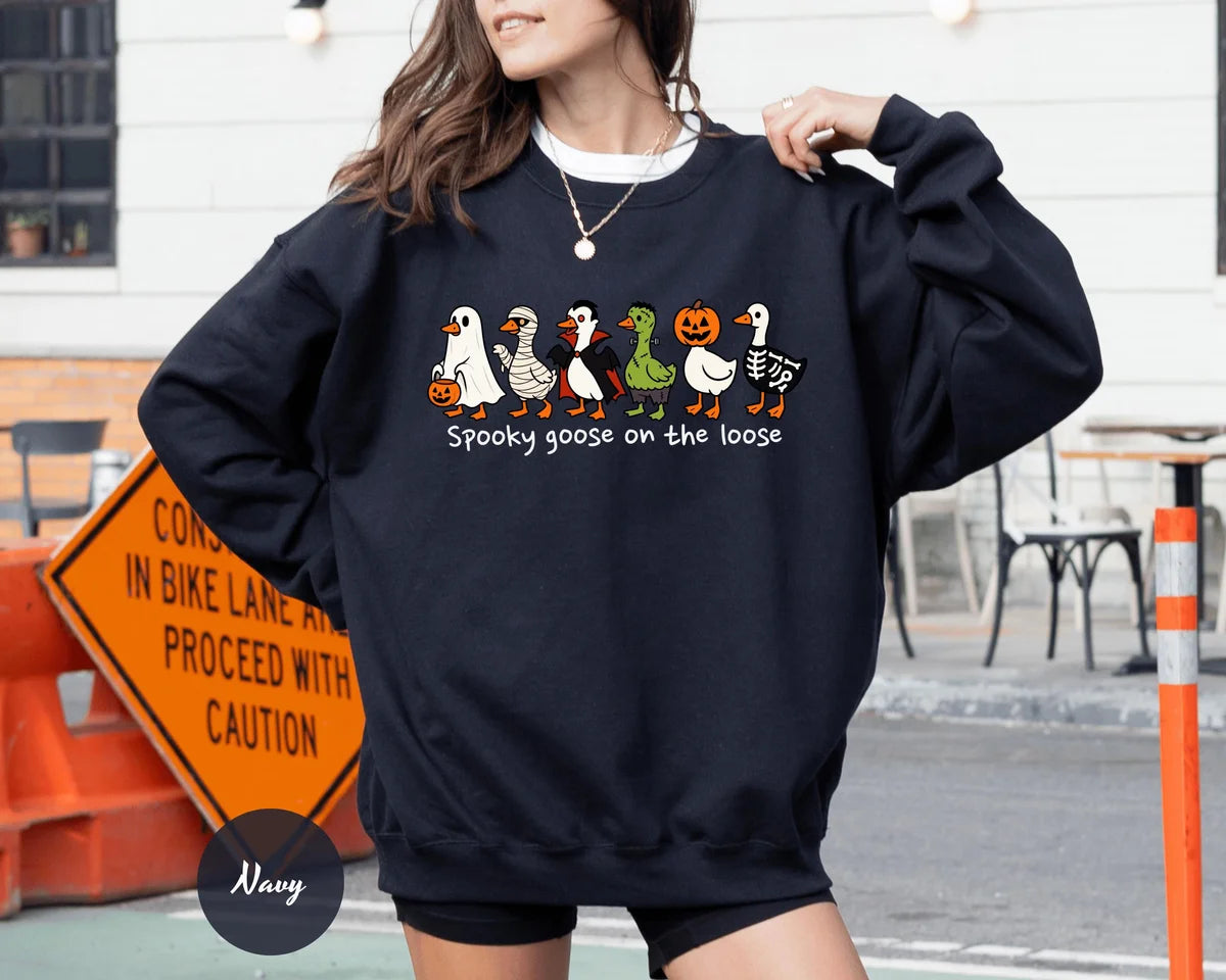 Navy crewneck sweatshirt with costumed geese dressed as a ghost, vampire, pumpkin and skeleton above the words spooky goose on the loose, worn by a model.