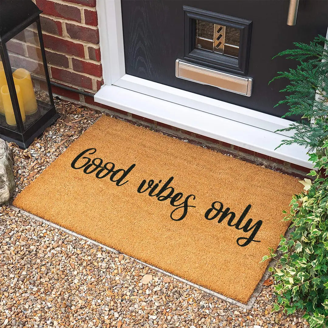 Rectangular coir doormat with Good Vibes Only cursive lettering on gravel in front of a black door