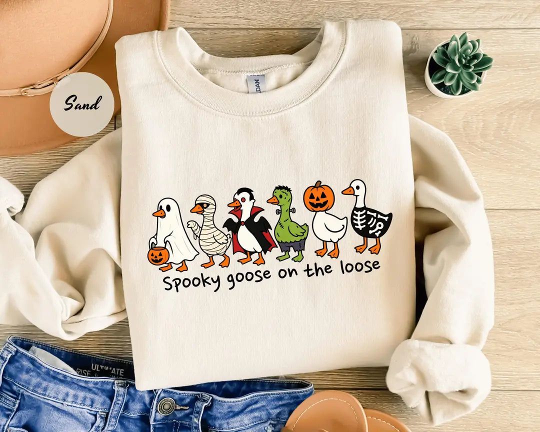 Sand crewneck sweatshirt with costumed geese dressed as a ghost, vampire, pumpkin and skeleton above the words spooky goose on the loose, flat lay view.