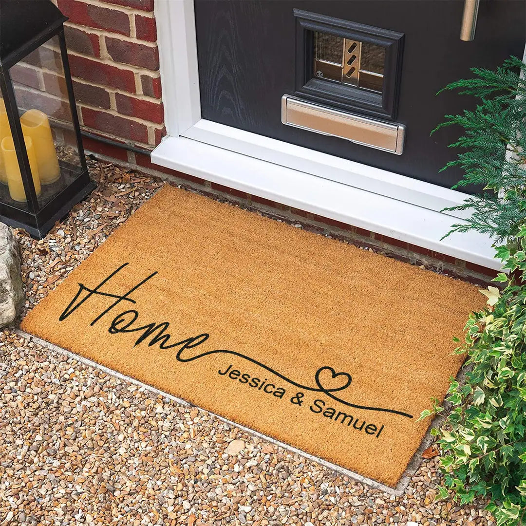 Rectangular coir doormat with a heart script Home and two names on gravel in front of a black door