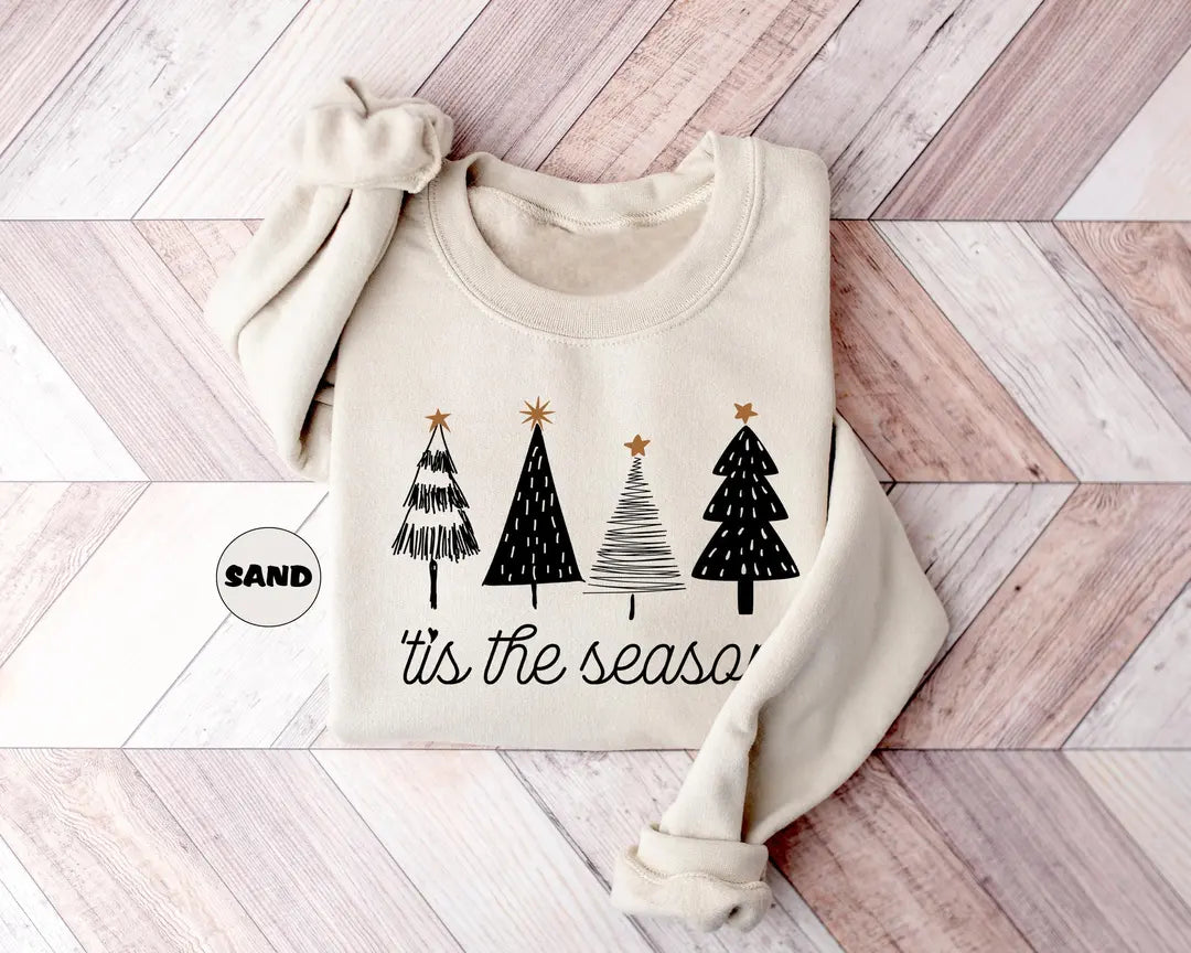 Sand crewneck sweatshirt with four sketch-style Christmas trees above the script words tis the season, flat lay view.