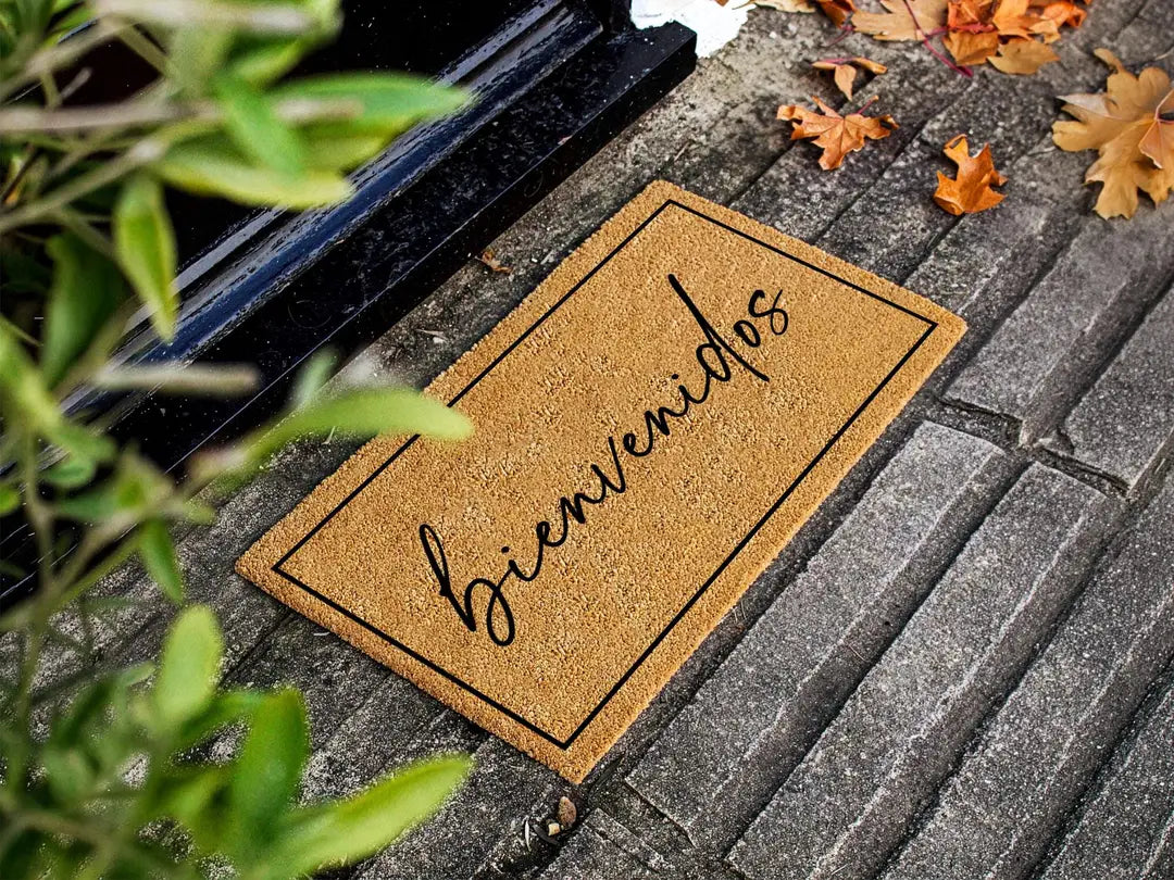 Coir doormat with a framed bienvenidos script message on dark stone steps with autumn leaves and green foliage
