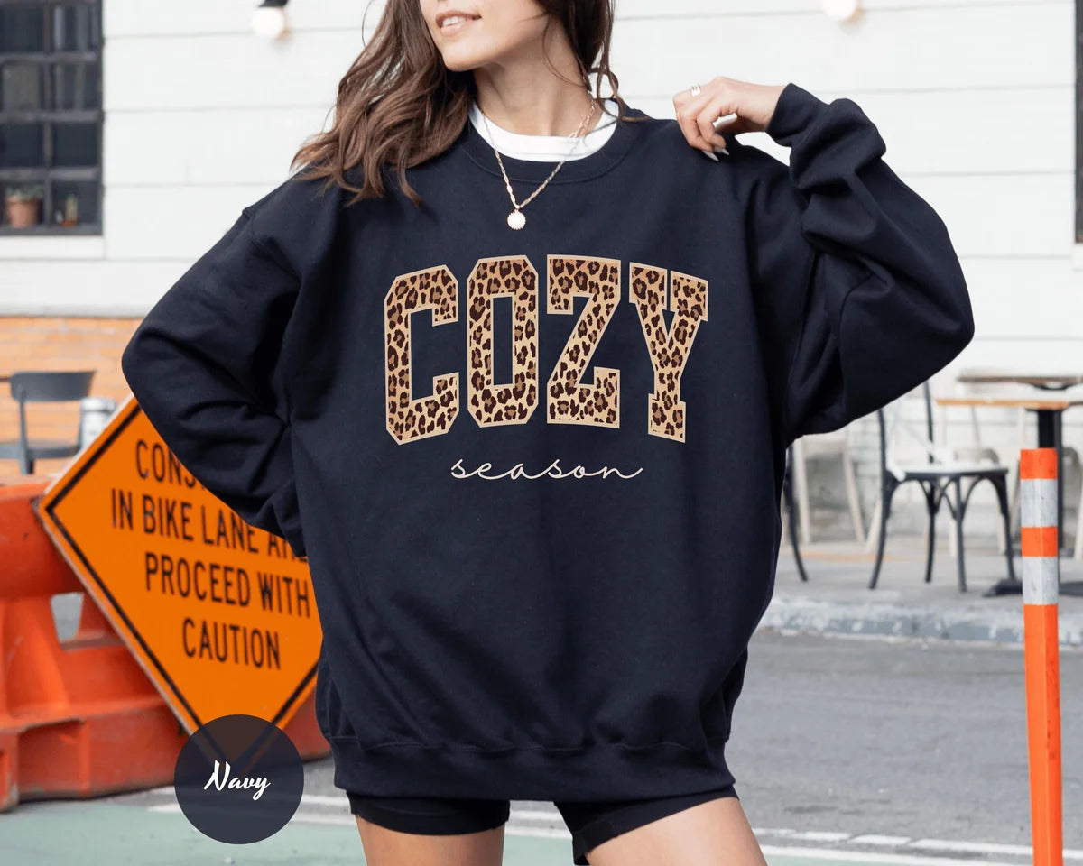 Navy crewneck sweatshirt with bold leopard print cozy lettering above the script words season, worn by a model.