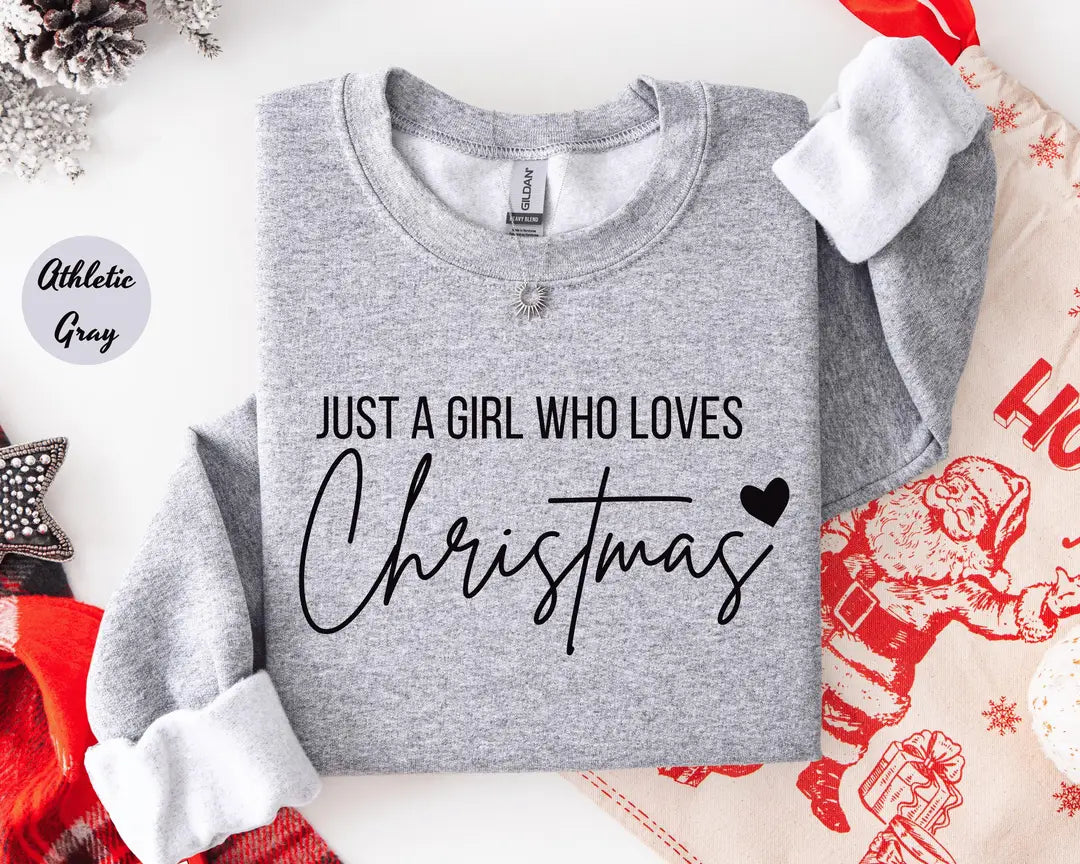 Athletic Gray crewneck sweatshirt with script lettering that reads just a girl who loves Christmas with a small heart, flat lay view.