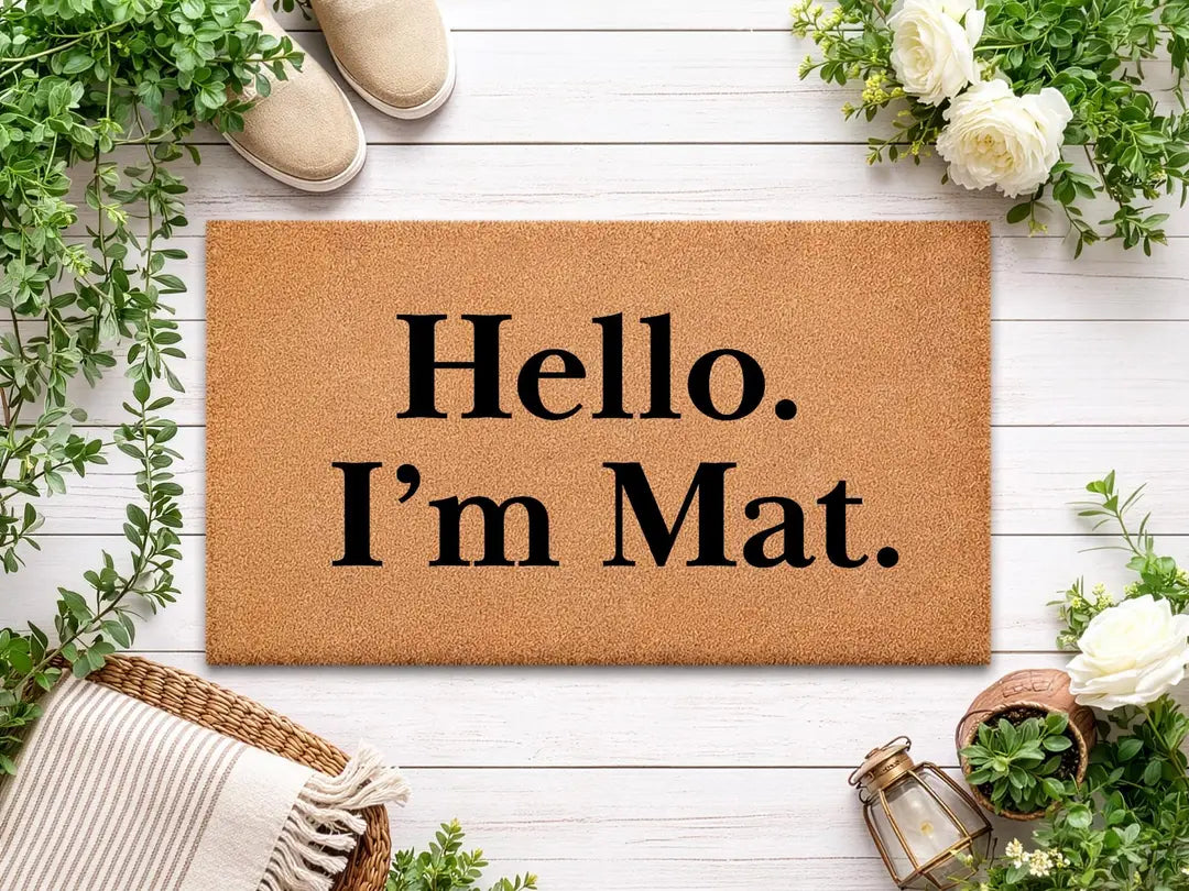 Coir doormat with a Hello I'm Mat message on white wood boards with greenery, white roses, and a woven basket