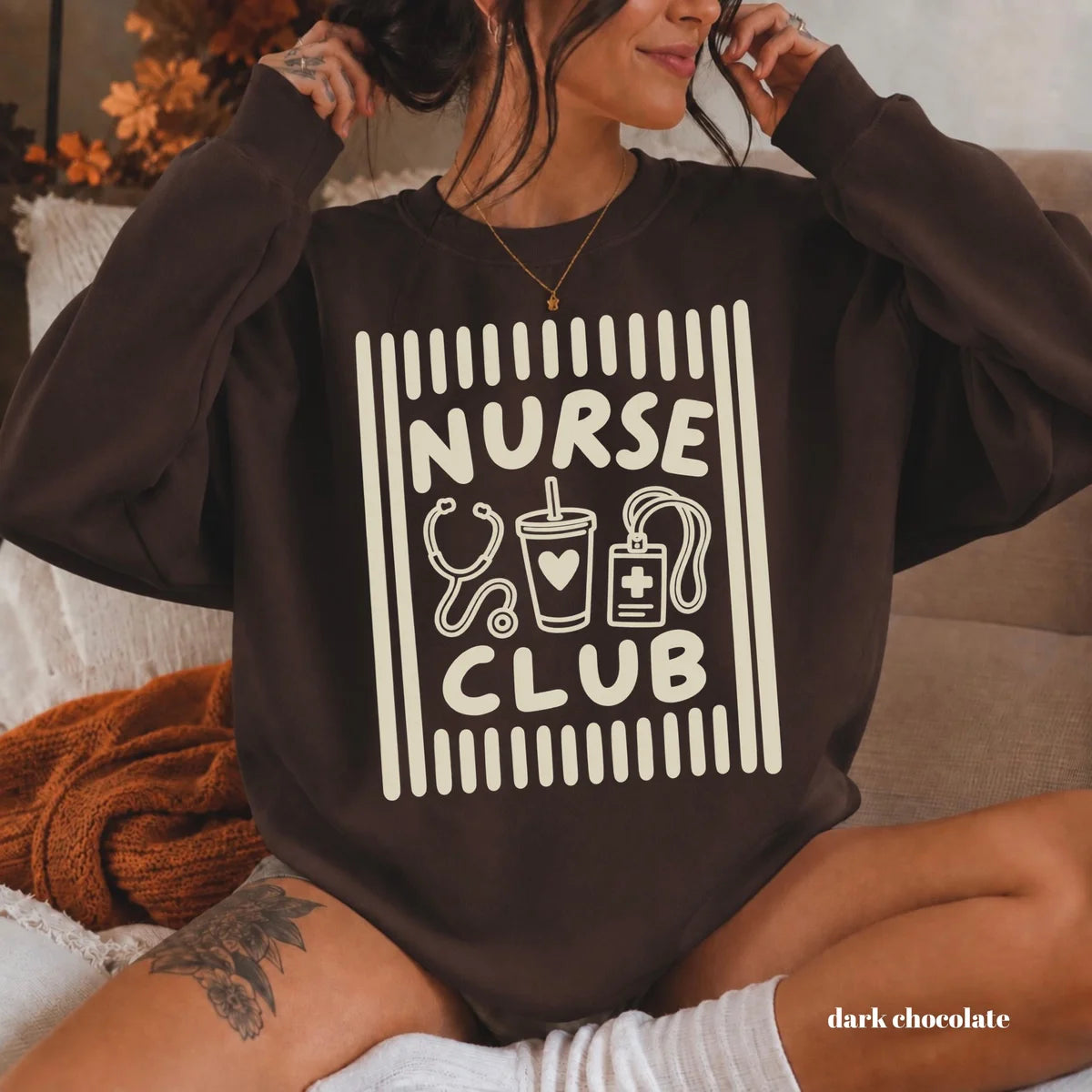 Dark Chocolate crewneck sweatshirt with printed a retro striped frame with a stethoscope, coffee cup and badge above the words nurse club, worn by a model.