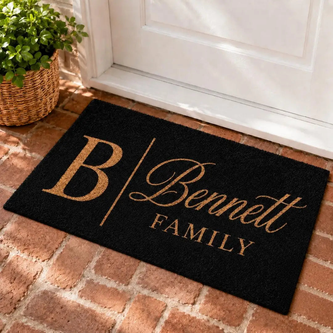 Rectangular black faux coir doormat with a gold-tone monogram initial, script surname, and Family line at a white door on brick