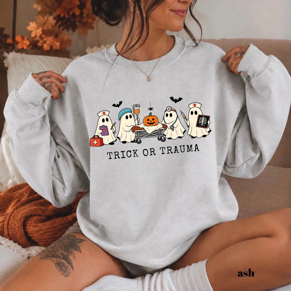 Ash crewneck sweatshirt with printed nurse ghosts with a pumpkin and the words trick or trauma, worn by a model.