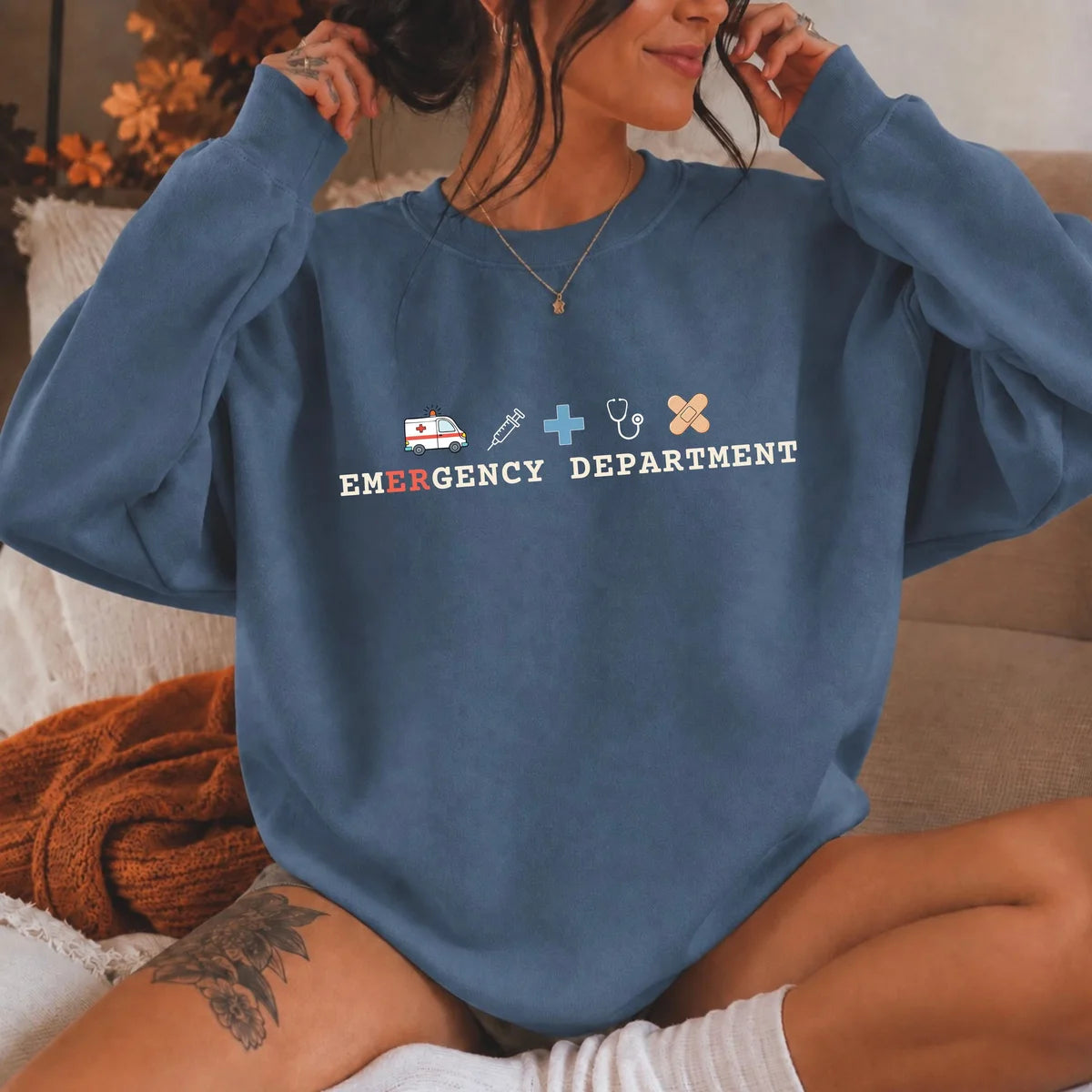 Indigo Blue crewneck sweatshirt with printed an ambulance, syringe, cross, stethoscope and bandage above the words emergency department, worn by a model.