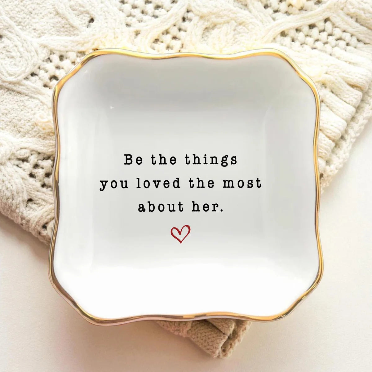 Square ceramic memorial ring dish with gold rim resting on a cream knit blanket