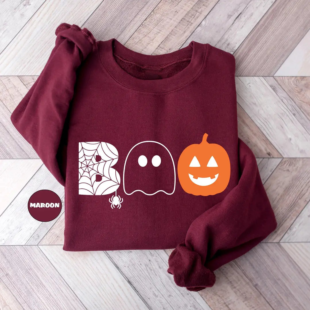 Maroon crewneck sweatshirt with the word boo spelled with a spider web letter, a ghost and a jack-o-lantern, flat lay view.