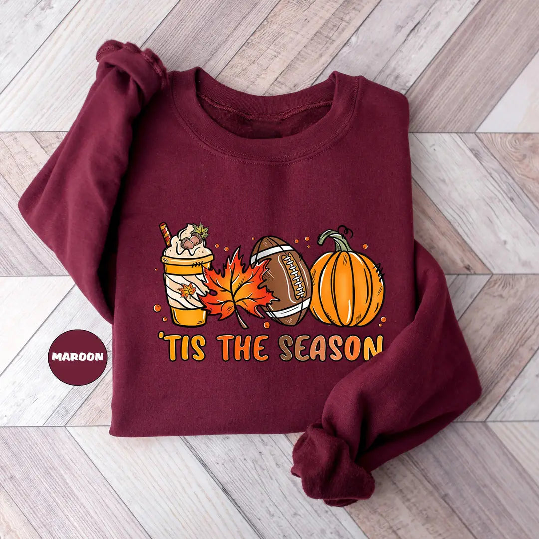 Maroon crewneck sweatshirt with a latte, autumn leaves, a football and a pumpkin above the words tis the season, flat lay view.