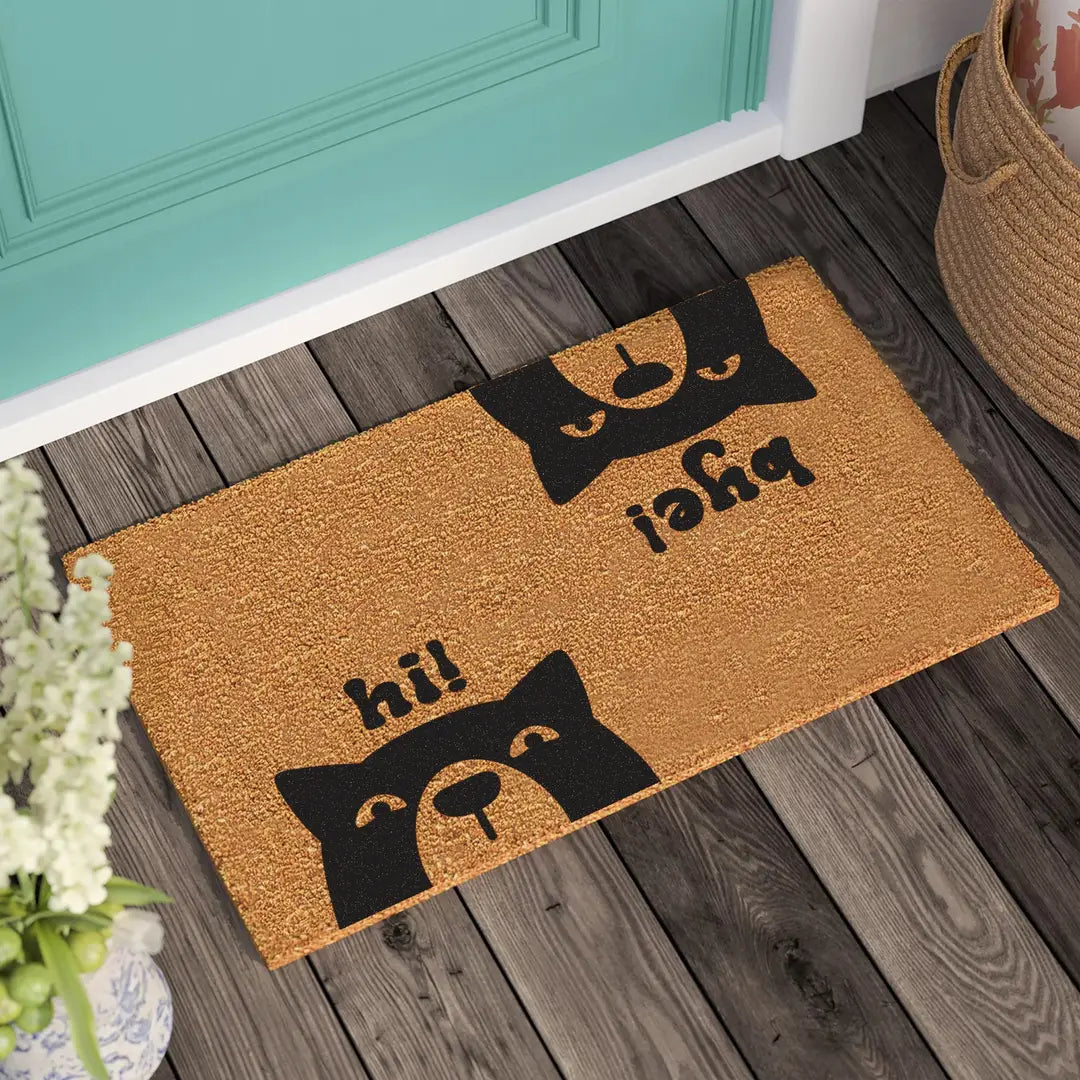 Coir doormat with two dog faces and hi and bye lettering on a wooden deck in front of a teal door