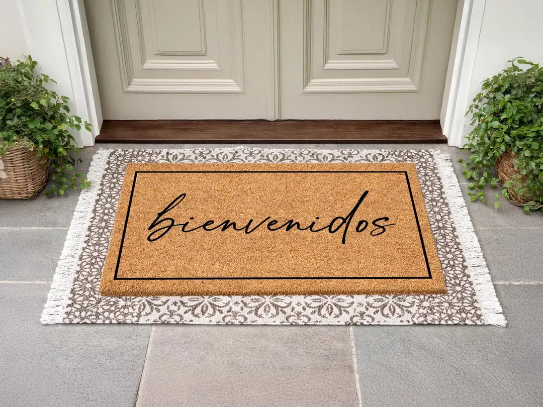 Rectangular coir doormat with a framed bienvenidos script message layered on a gray patterned fringed rug at a cream door