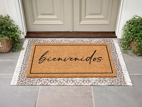 Rectangular coir doormat with a framed bienvenidos script message layered on a gray patterned fringed rug at a cream door