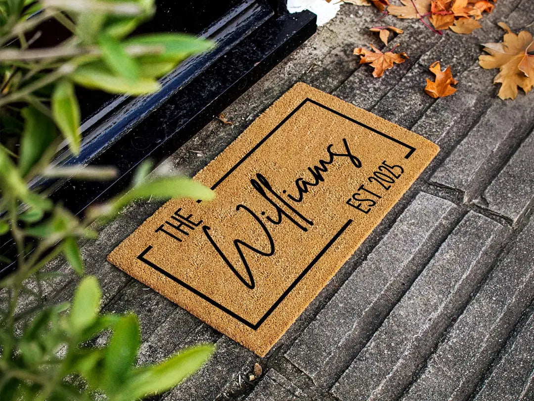 Coir doormat with a framed family name and established year on dark stone steps with autumn leaves and green foliage