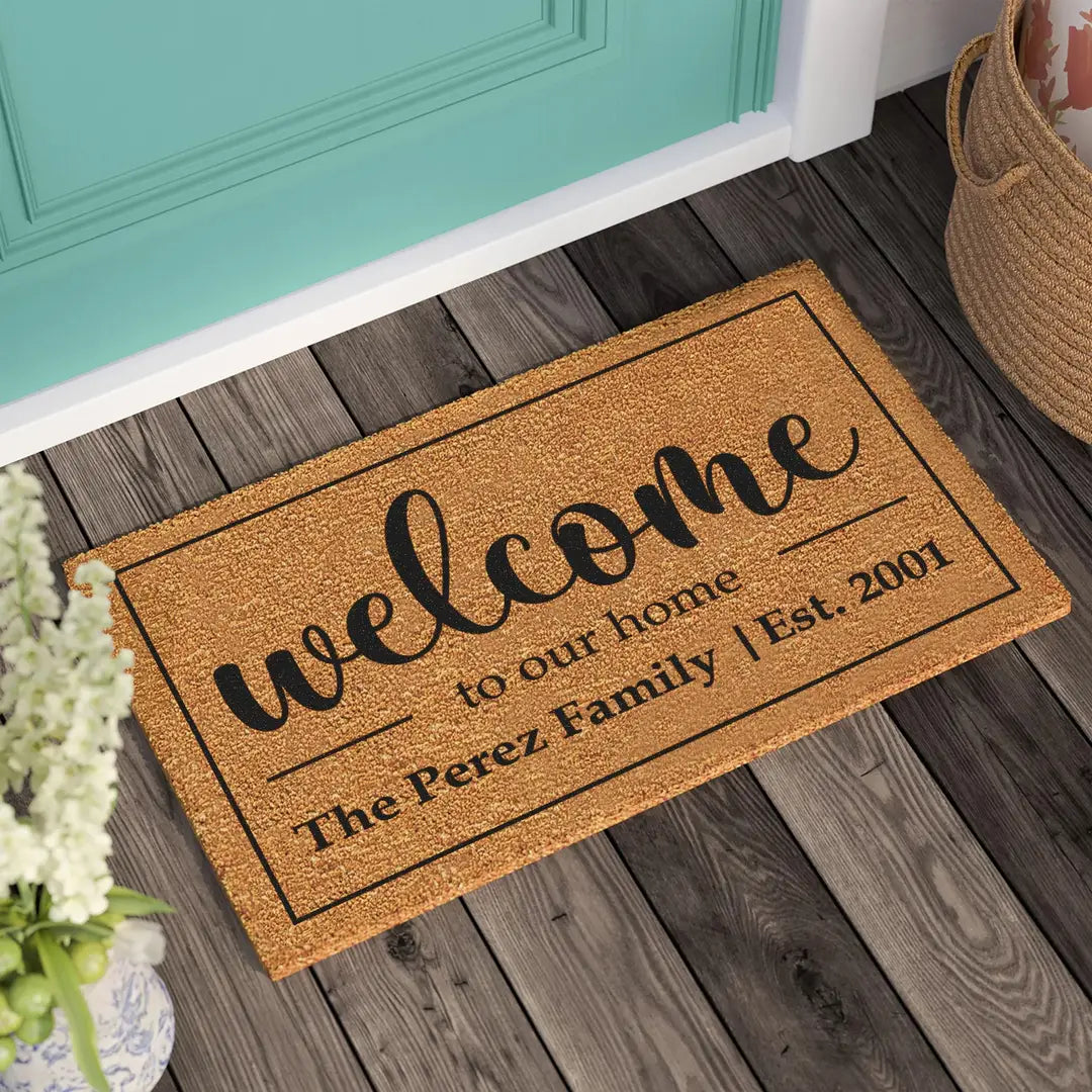 Coir doormat with a bordered welcome to our home design and family name on a wooden deck in front of a teal door