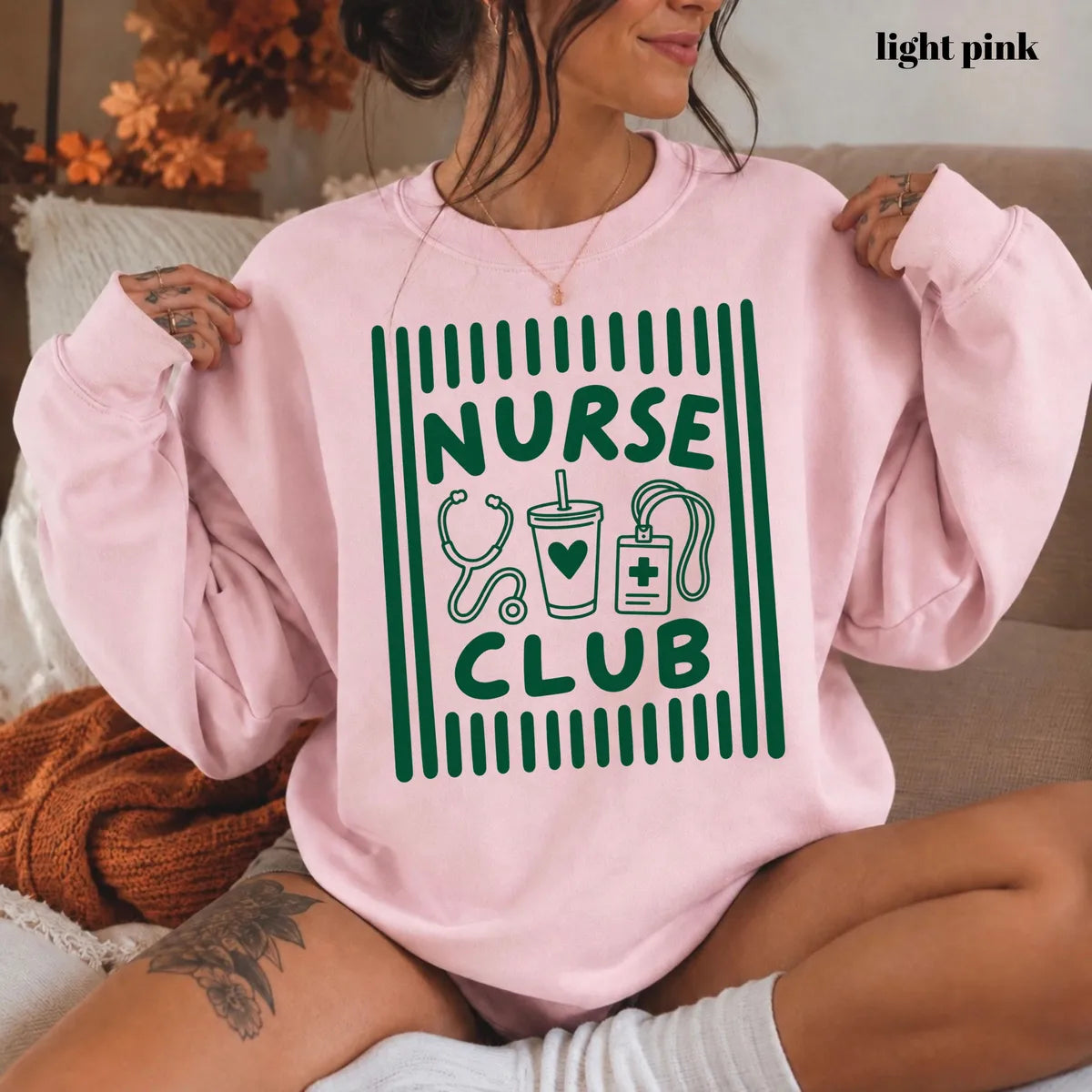 Light Pink crewneck sweatshirt with printed a retro striped frame with a stethoscope, coffee cup and badge above the words nurse club, worn by a model.