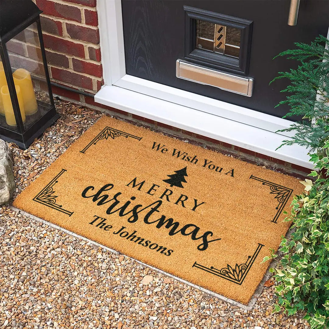 Rectangular coir doormat with a We Wish You a Merry Christmas message, pine tree, and family name on gravel in front of a black door