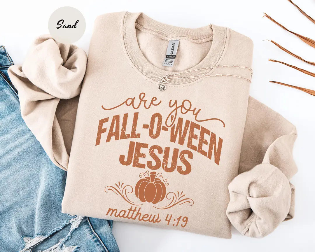 Sand crewneck sweatshirt with printed a pumpkin with the words are you fall-o-ween jesus and matthew 4:19, flat lay view.