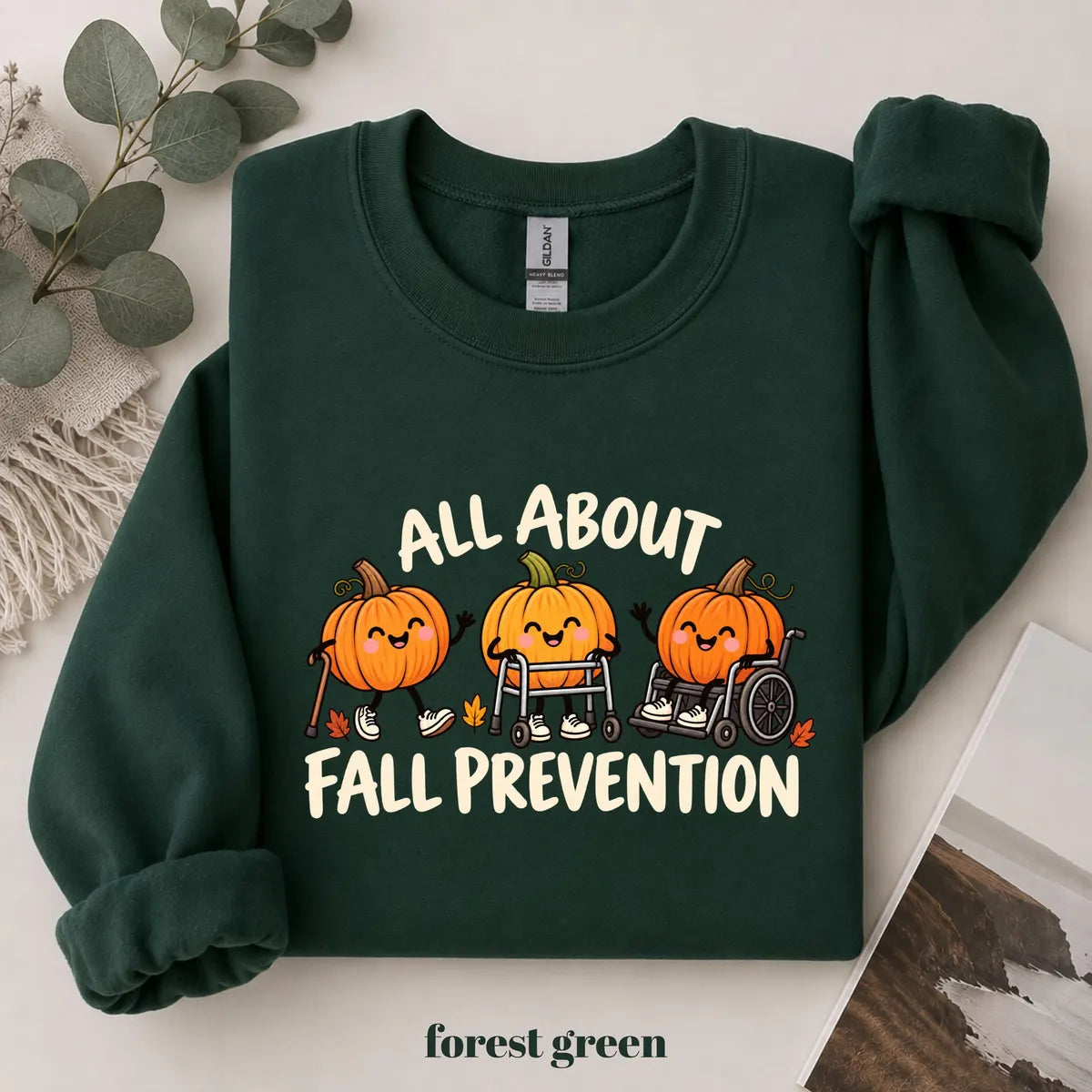 Forest Green crewneck sweatshirt with printed pumpkins with a walker and wheelchair above the words all about fall prevention, flat lay view.