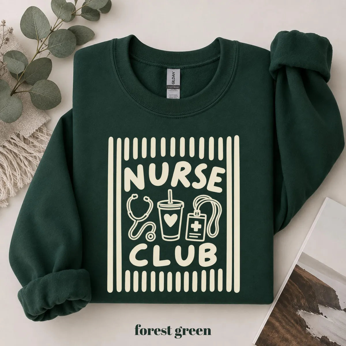Forest Green crewneck sweatshirt with printed a retro striped frame with a stethoscope, coffee cup and badge above the words nurse club, flat lay view.