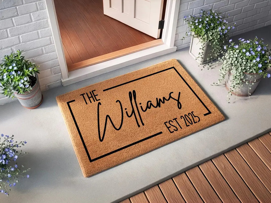Coir doormat with a framed family name and established year at an open white door with white brick siding and potted blue flowers