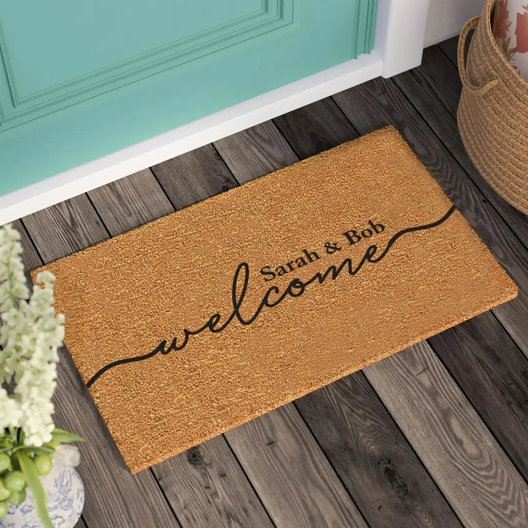 Rectangular coir doormat with flowing script welcome lettering and a couple's names in front of a teal door