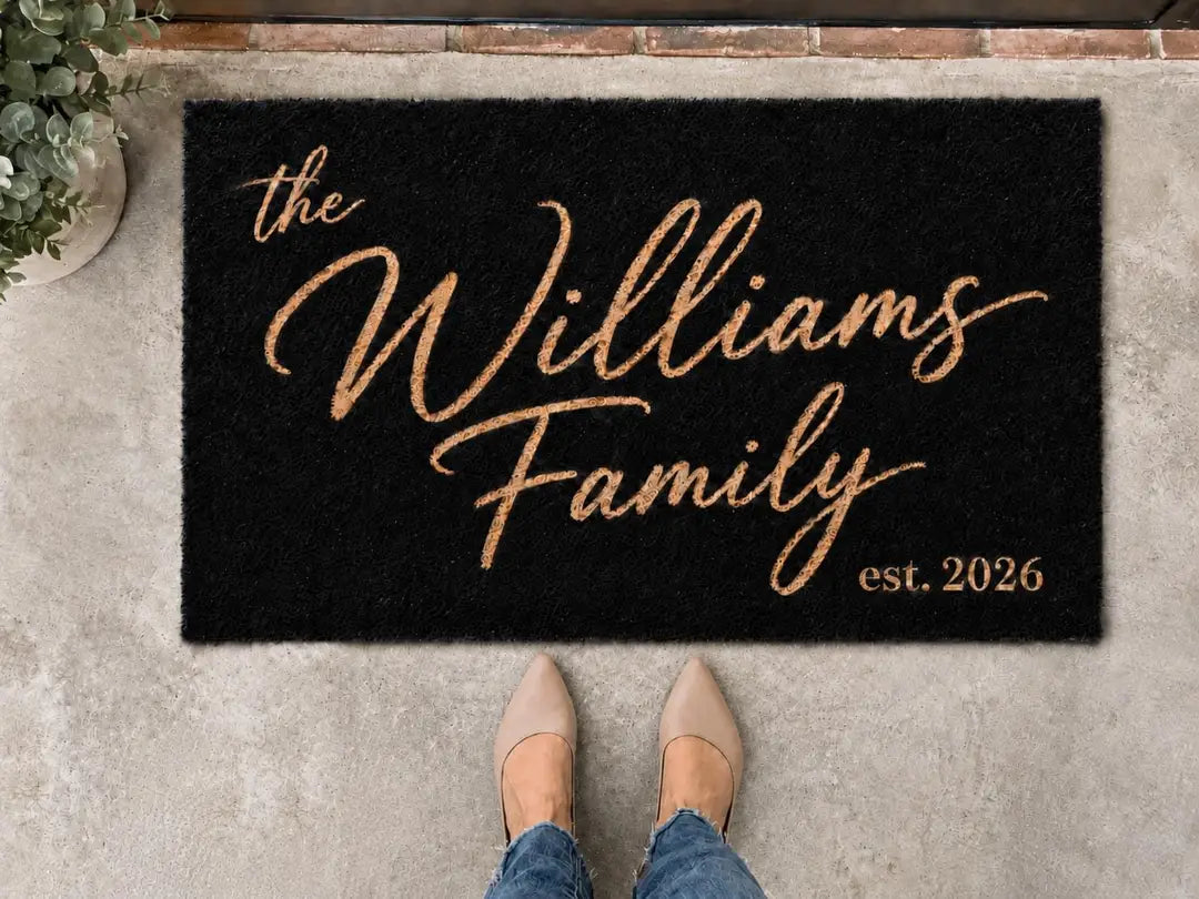 Black faux coir doormat with a gold-tone script family name and established year on a concrete step