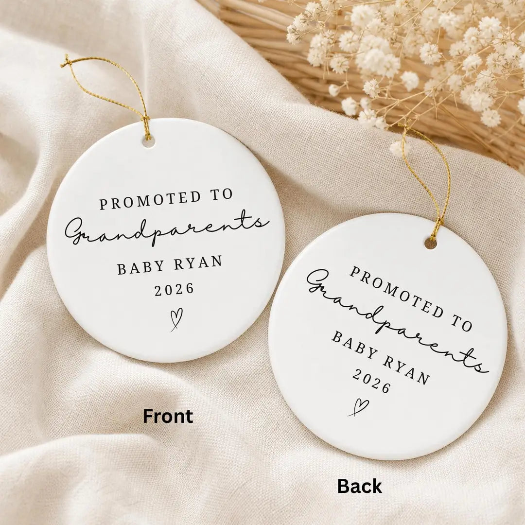 Front and back of a round white ceramic Promoted to Grandparents ornament with the same personalized baby name and year on both sides, beside dried flowers