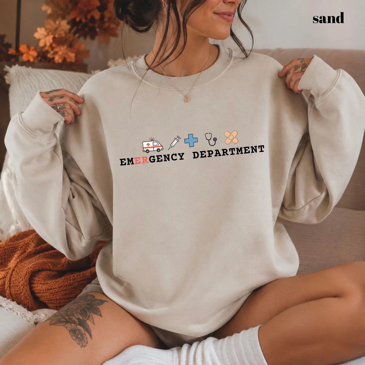 Sand crewneck sweatshirt with printed an ambulance, syringe, cross, stethoscope and bandage above the words emergency department, worn by a model.