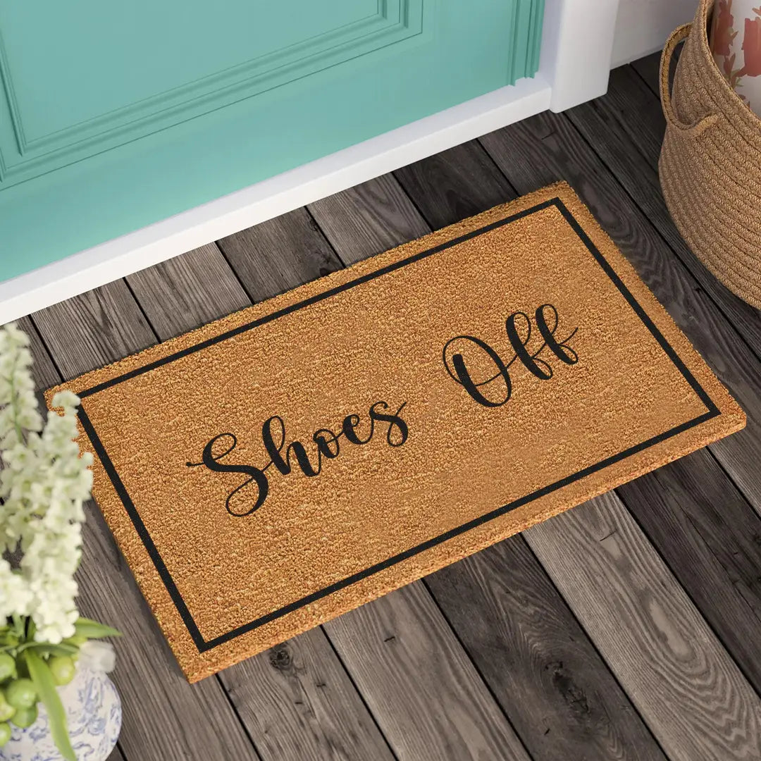 Coir doormat with Shoes Off script lettering and a black border on a wooden deck in front of a teal door