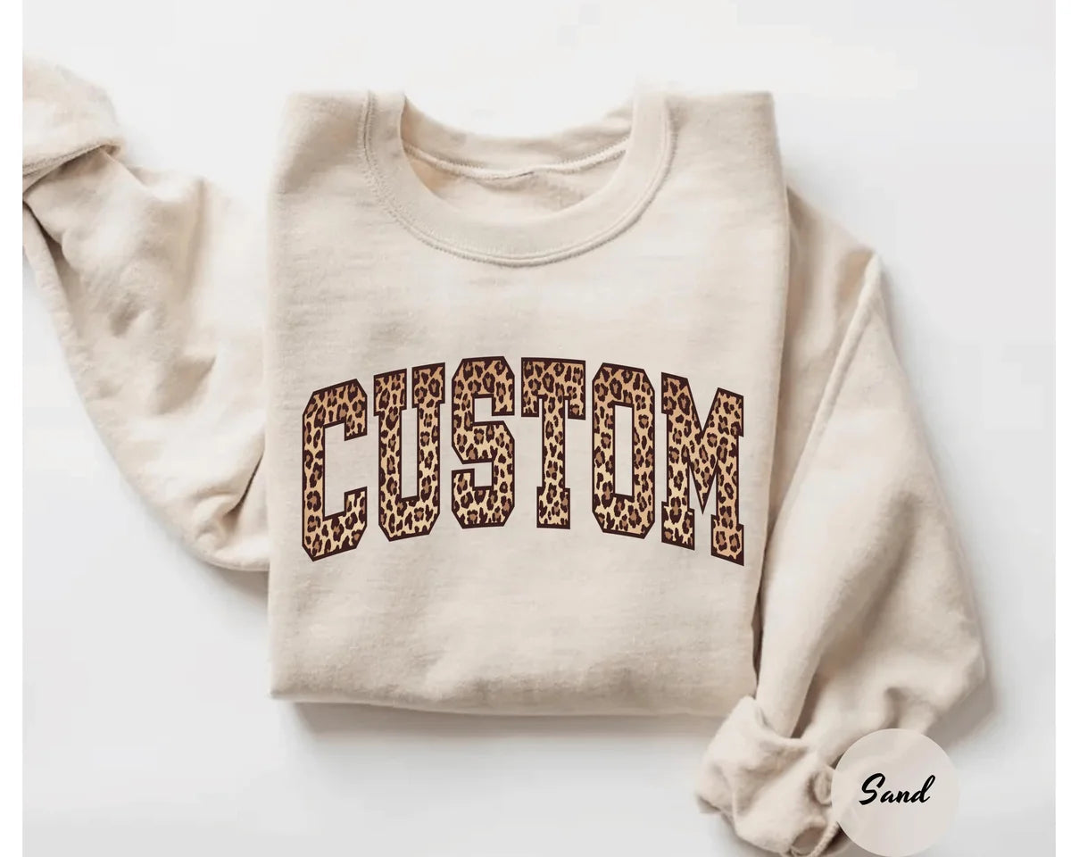 Sand crewneck sweatshirt with bold leopard print varsity lettering with your own custom name or text, flat lay view.