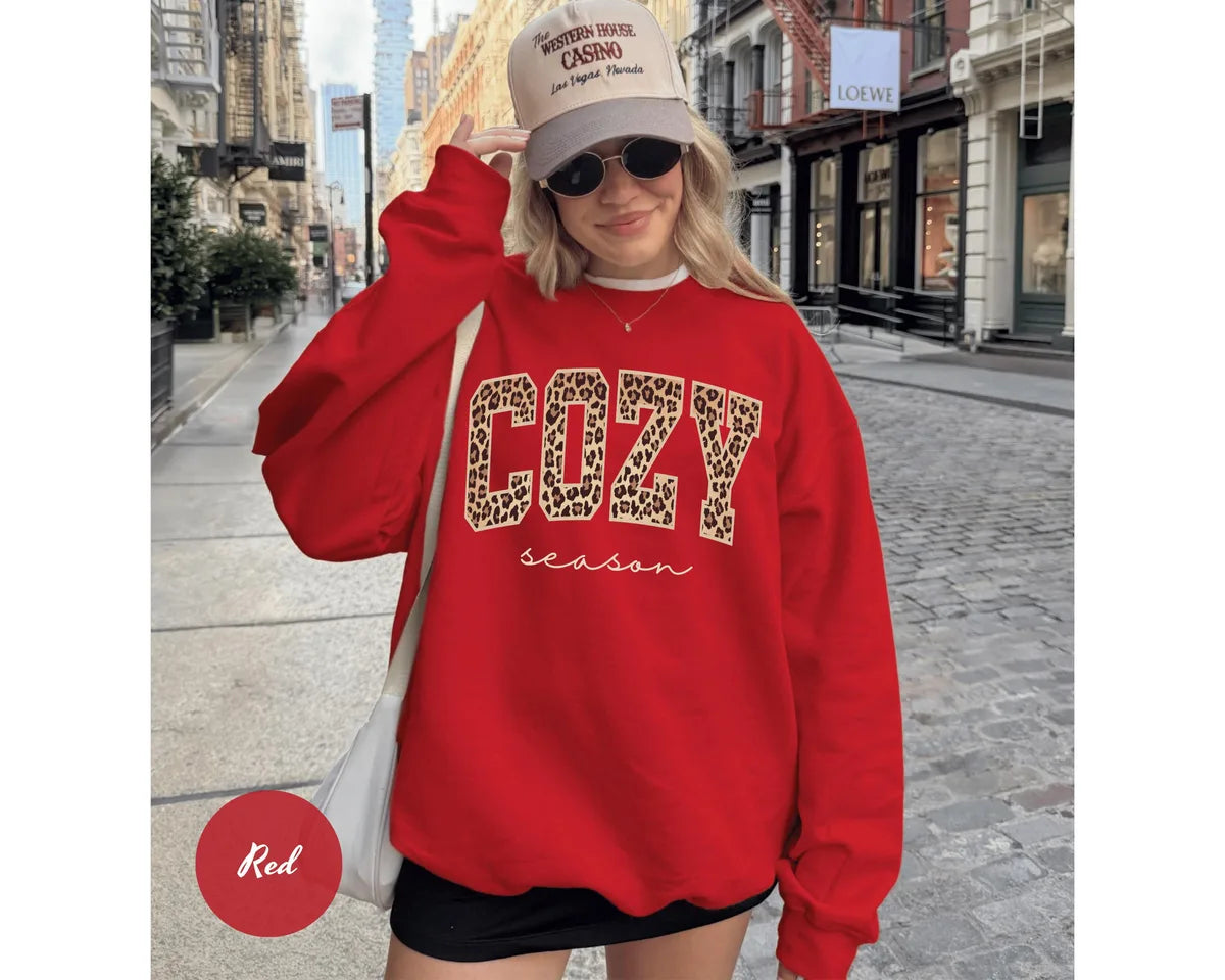 Red GD crewneck sweatshirt with bold leopard print cozy lettering above the script words season, worn by a model.